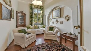 Ein geräumiges Wohnzimmer mit zwei weißen Sesseln, einem Sofa, einer Holzkommode und einem großen Fenster mit blumigen Vorhängen.