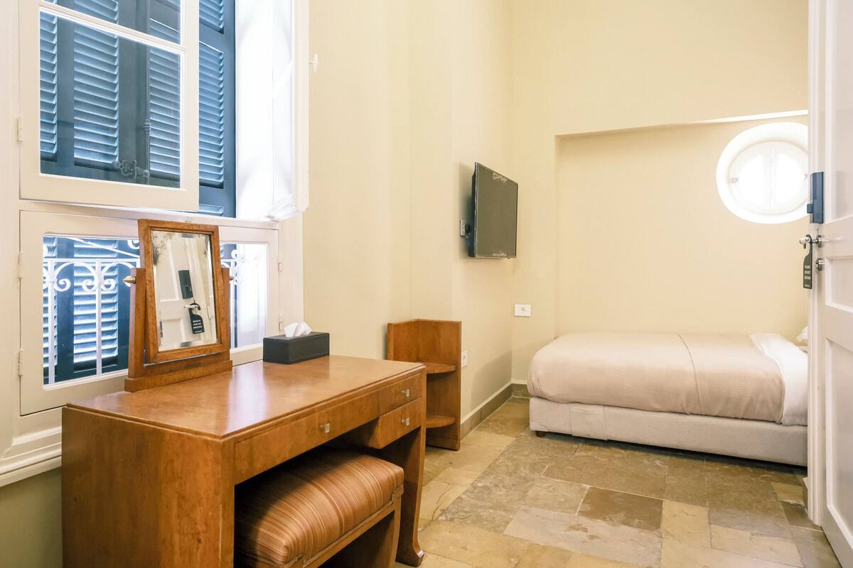 Un dormitorio con un escritorio de madera, un espejo, una mesita de noche, una cama y un televisor instalado en la pared.