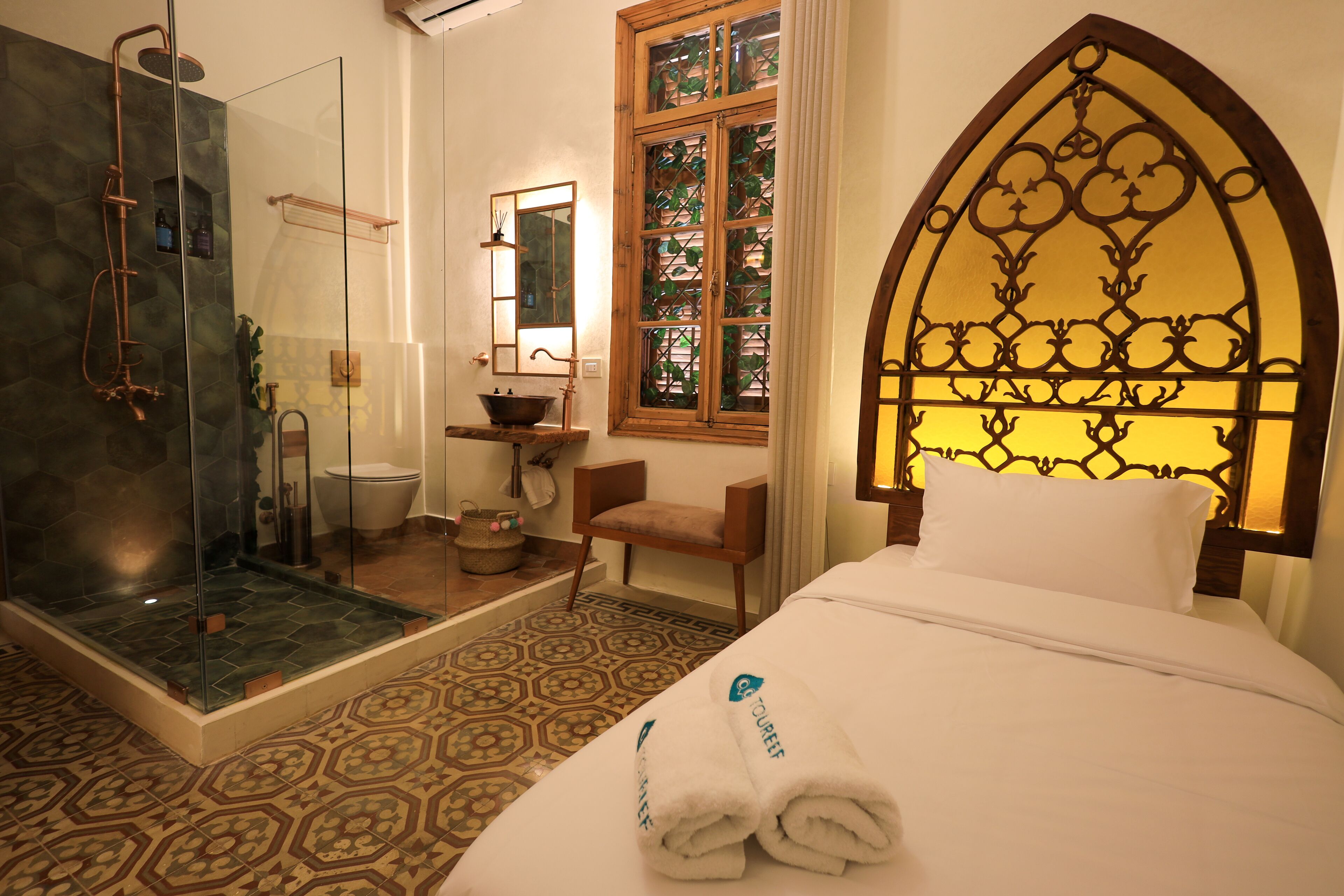 A hotel room with a shower, a sink, a chair, a bed with towels, a window with a decorative headboard, and a patterned floor.