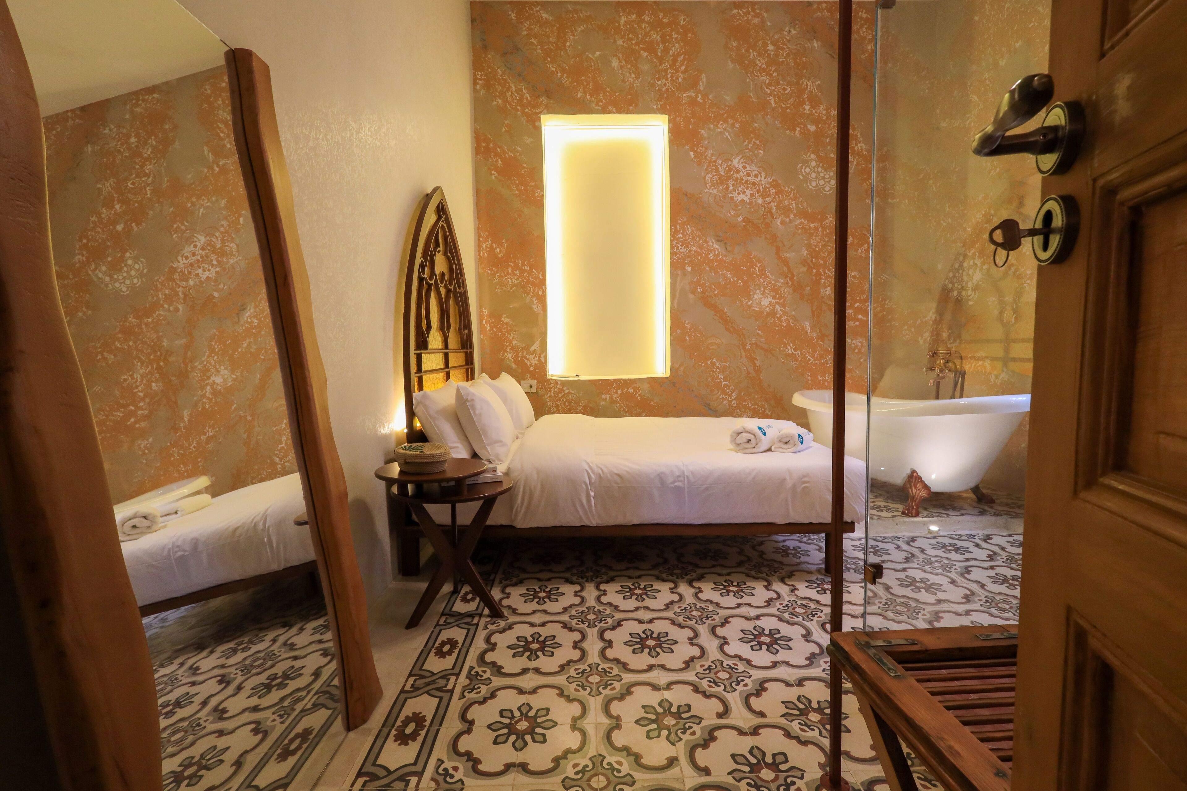 Un dormitorio con cama, mesita de noche y lámpara de noche. Hay una ventana con una luminaria sobre la cama.