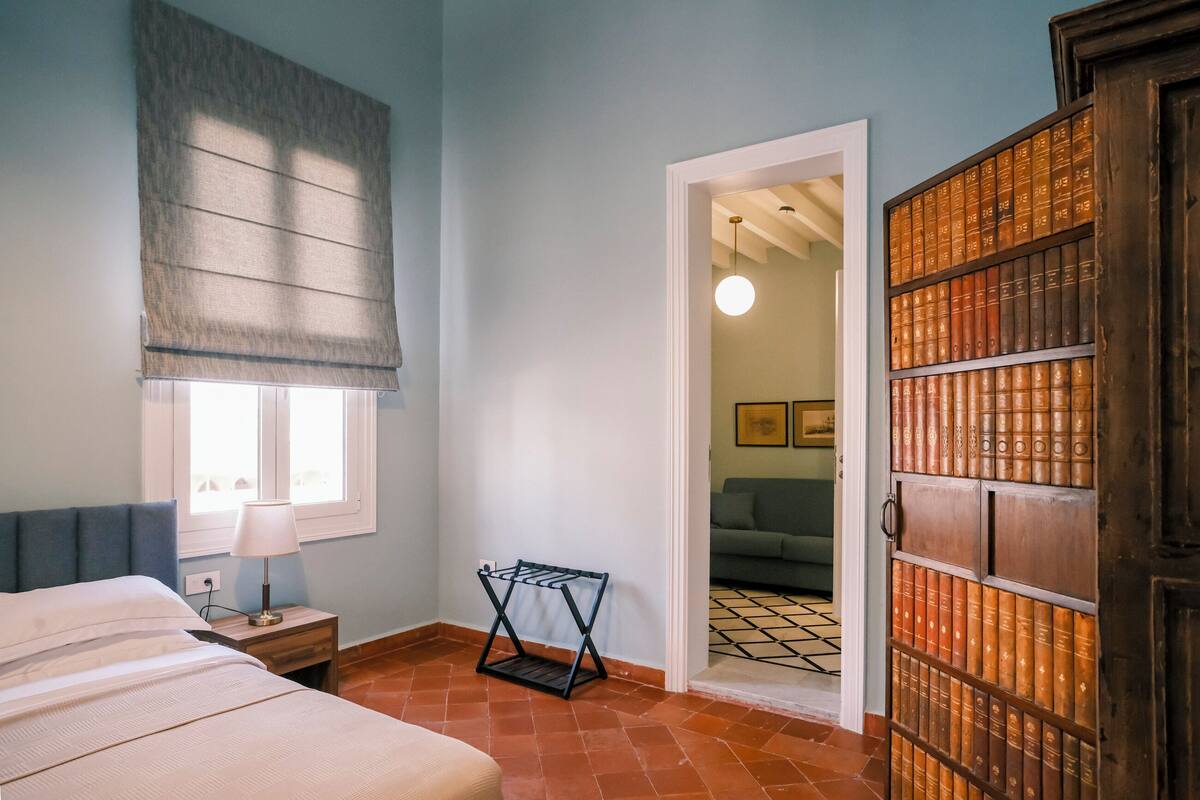 Un dormitorio con cama, mesita de noche, lámpara, ventana con persiana, puerta que da a otra habitación y estantería con libros.