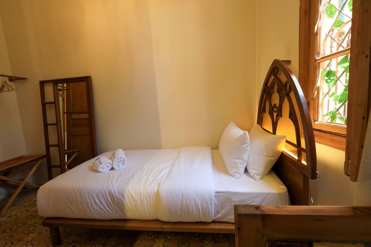 Una cama bien hecha con sábanas y almohadas blancas, un cabecero de madera y una ventana con vistas a la vegetación.