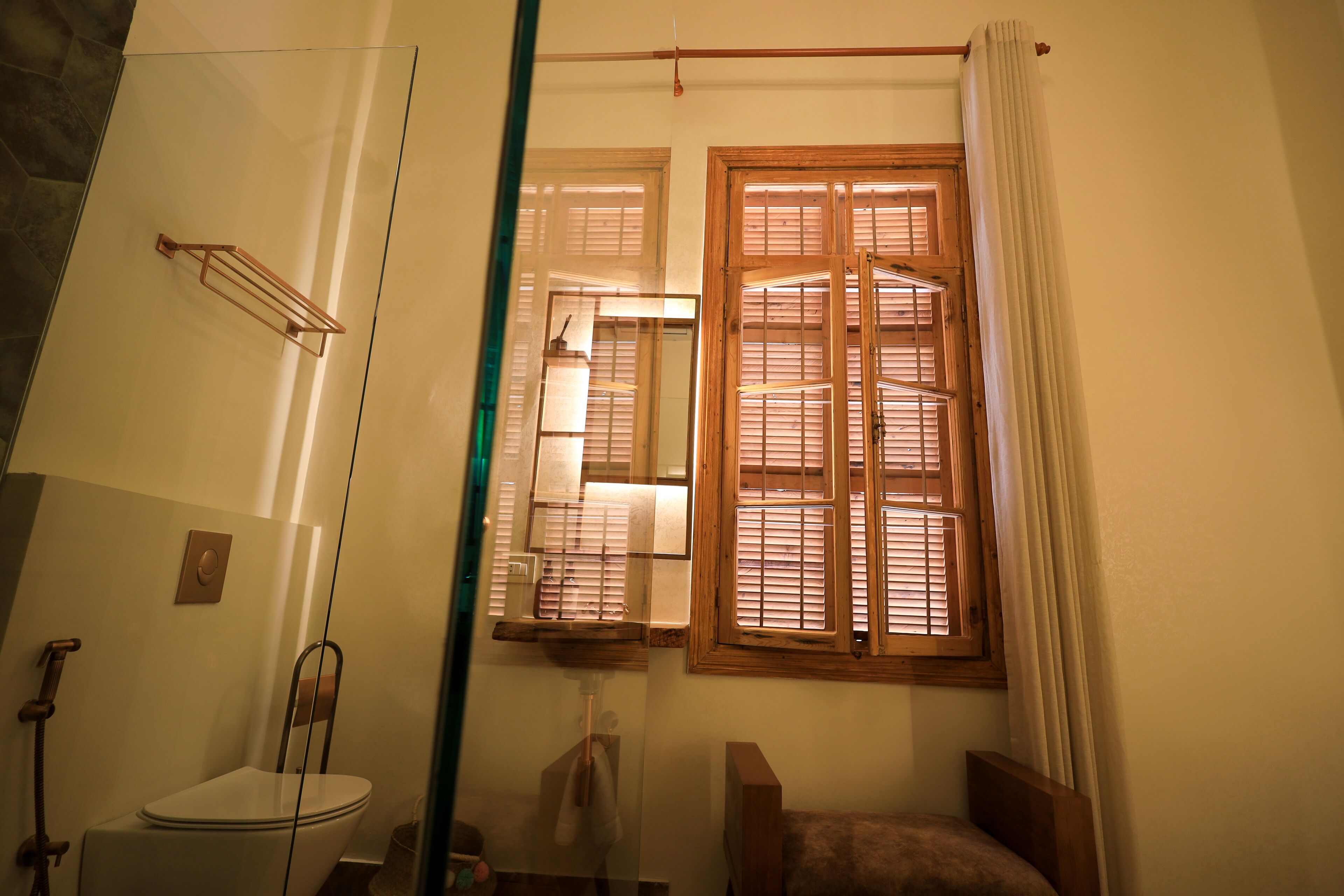 Baño con inodoro, lavamanos y zona de ducha. Hay una ventana con contraventanas de madera y una cortina.