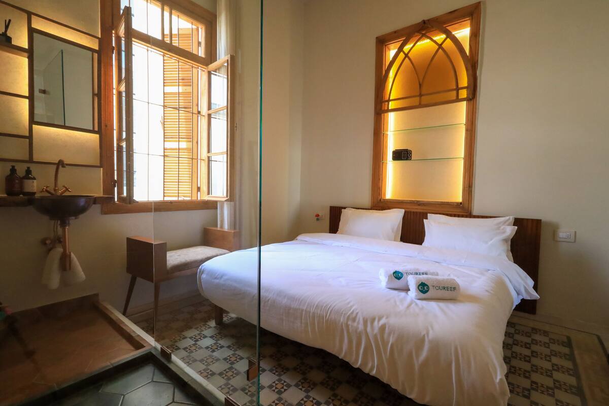Habitación de hotel con una cama grande, una mesita de noche, un espejo, una ventana con persianas y una pequeña estantería.