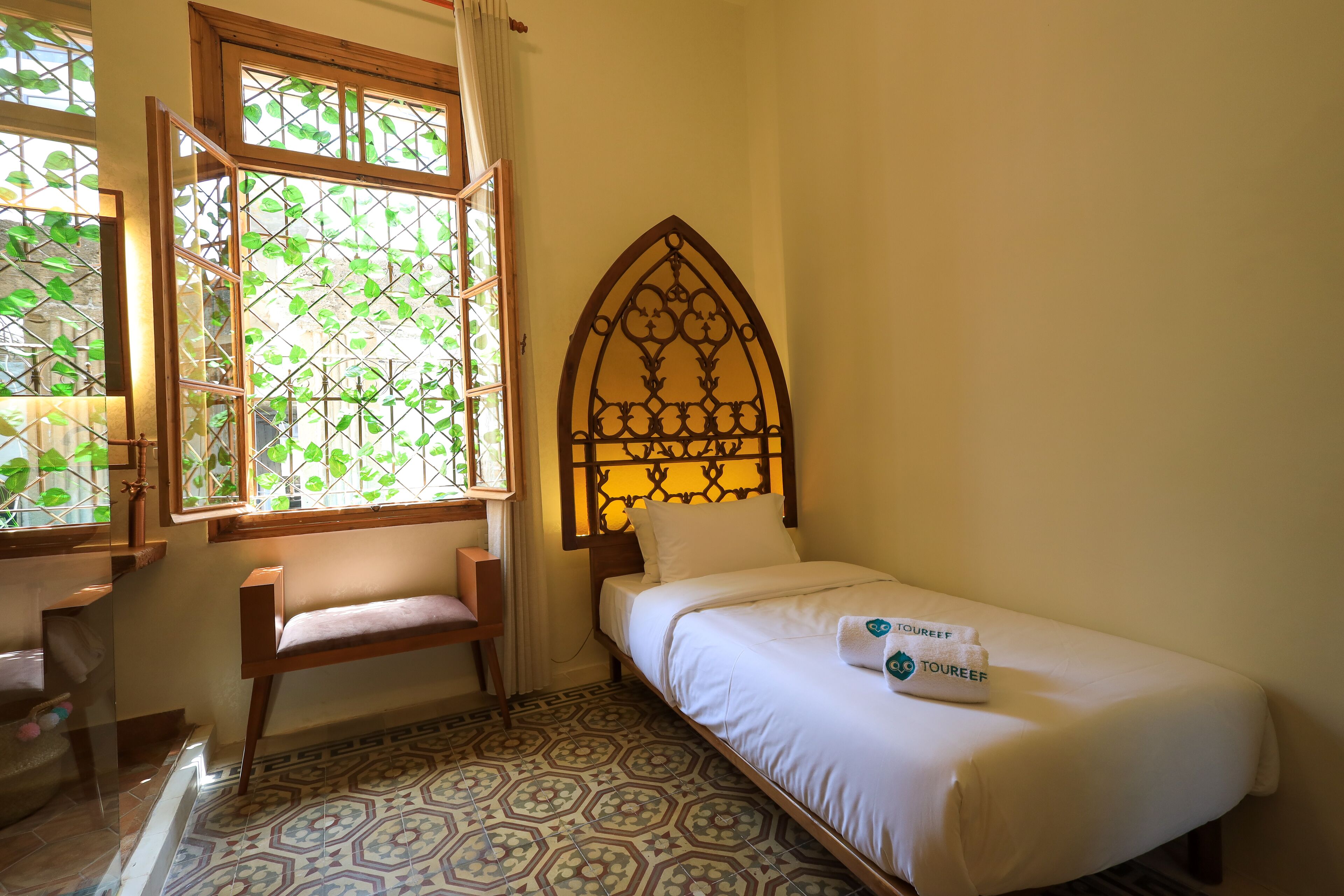 A single bed with a headboard, a chair, a window with a view of greenery, and patterned floor tiles.