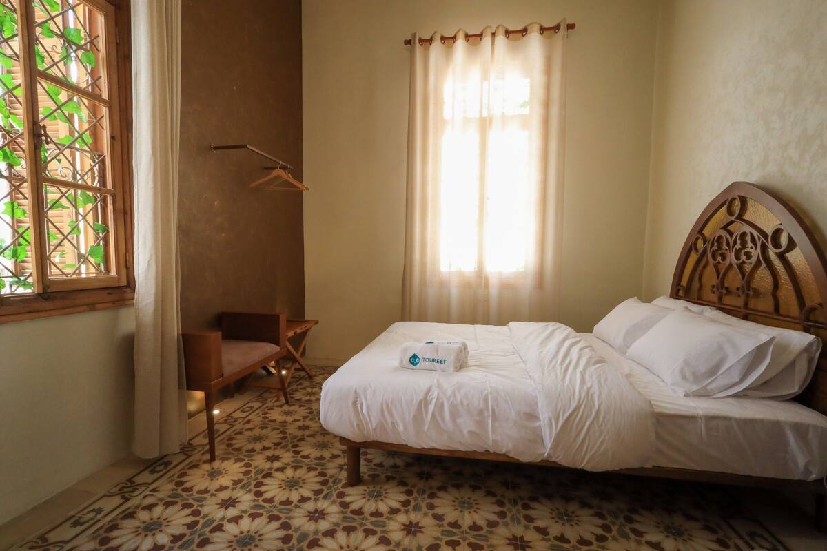 Una cama con cabecera, una silla, una ventana con cortina y un piso con diseño.