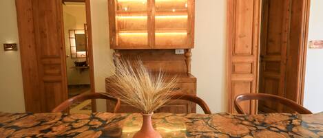 A dining table with a vase of dried wheat stalks, wooden chairs, and a wooden cabinet with a lighted panel in the background.