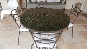 Een ronde tafel met een mozaïekte top, omringd door vier stoelen, een bank met kussens en een witte stoel in een kamer met betegelde vloer.