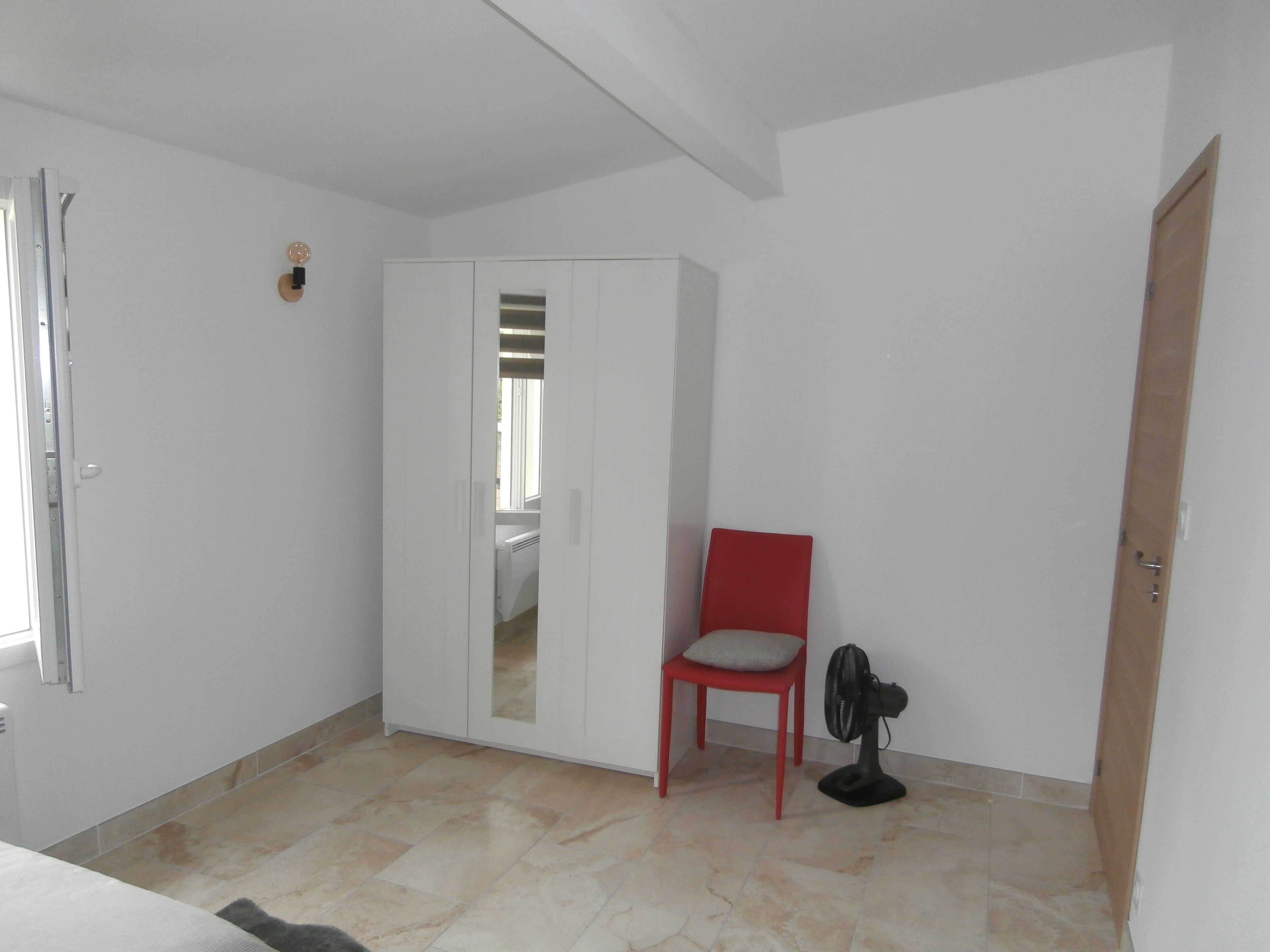 Una habitació amb un armari blanc, una cadira vermella, un ventilador de peu i un terra solcat.