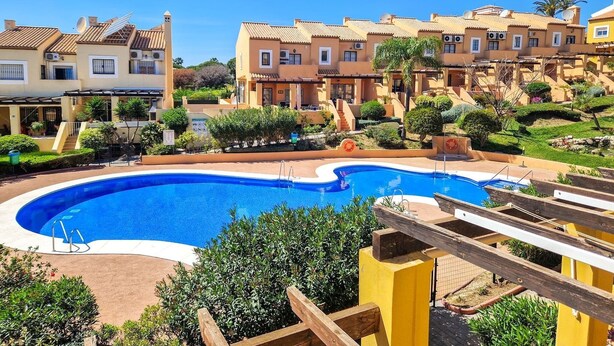 A swimming pool surrounded by terraced buildings with balconies, a clear blue sky, and lush greenery.