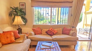 A living room with a beige sofa, a coffee table with fruits, a lamp, and a view of greenery outside.