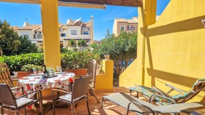 A patio with a table set for dining, surrounded by wicker chairs and lounge chairs, overlooking a residential area with buildings and greenery.