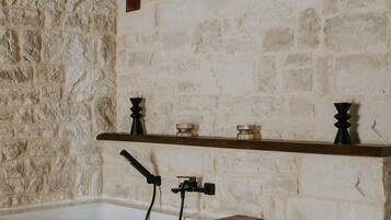 Un baño con pared de piedra, repisa de madera con objetos decorativos y una bañera con grifo.