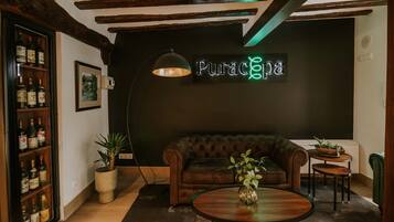 Una habitación acogedora con un sofá de cuero, una mesa de centro redonda y una estantería llena de botellas. Un letrero de neón dice "Puracpa" en la pared.