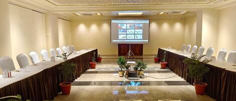 Un salón de conferencias con una larga mesa, sillas blancas y una pantalla proyectando una presentación.