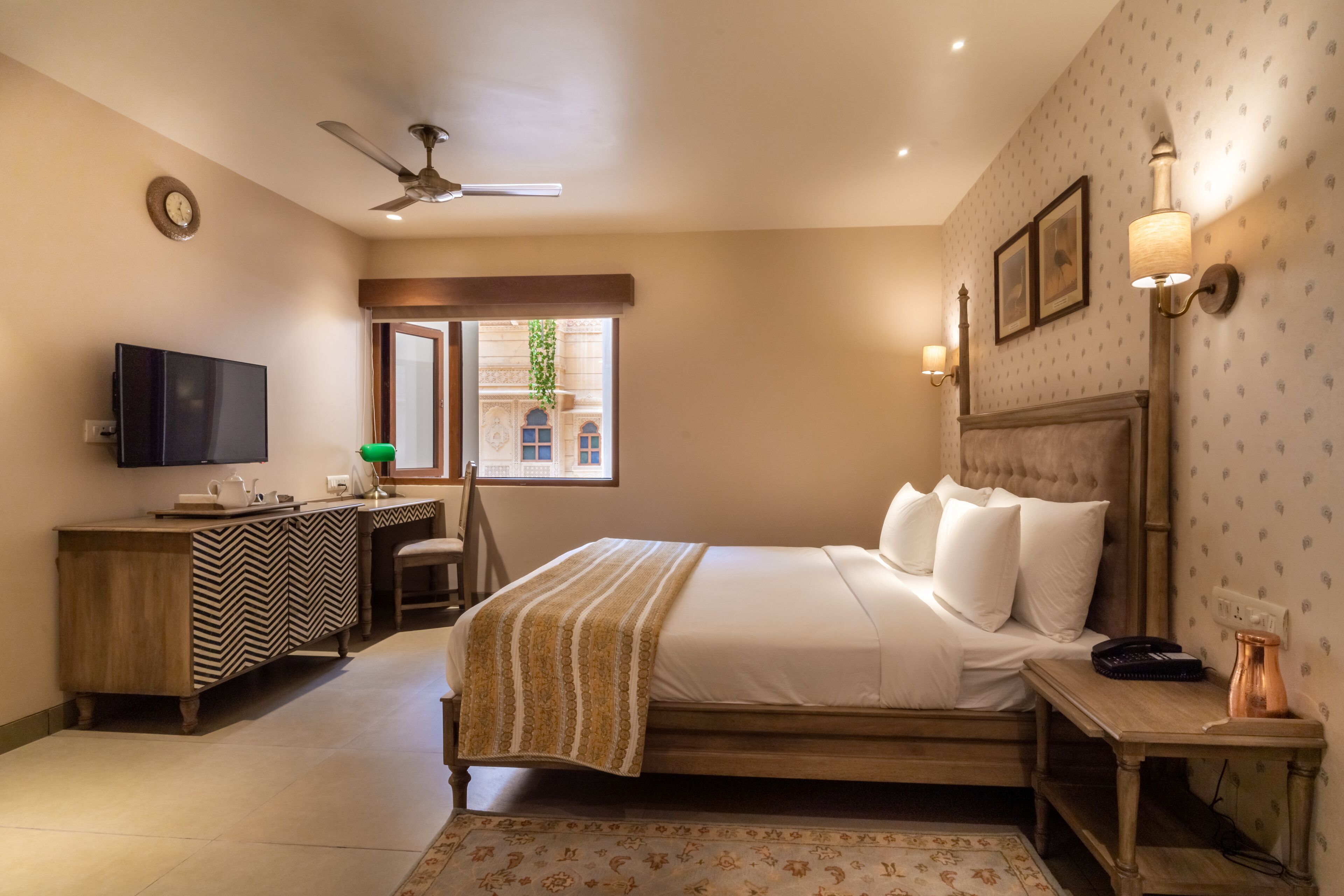 A hotel room with a bed, a desk, a television, a ceiling fan, and a window with a view of buildings.