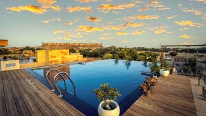 Una piscina infinita en la azotea, con cubierta de madera, plantas en macetas y vista a un horizonte urbano distante.