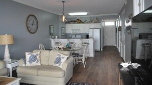Una sala de estar con sofá, una mesa de comedor con sillas, una cocina con armarios y electrodomésticos, y un pasillo que conduce a una puerta.