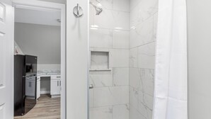 A modern bathroom with white marble tiles, a glass shower enclosure, and a visible kitchen area with white cabinets and a black refrigerator. - Cozy & Quiet Studio Flat in Historic Mount Clemens (Mount Clemens)