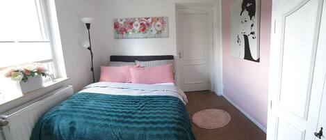 A bedroom with a bed, a radiator, a window, a door, and a wall with a painting.