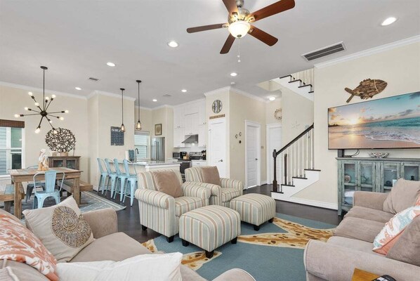 A spacious living room with striped sofas, a dining area with blue chairs, a kitchen, and a staircase. - Relaxing 4BR Beachside Home with Hot Tub | Pet Friendly (Galveston)