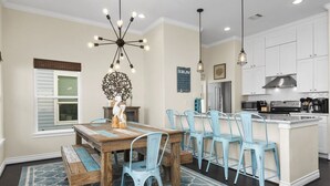 Un comedor con una mesa de madera y sillas azules, un candelabro, y una cocina con armarios blancos y electrodomésticos de acero inoxidable.