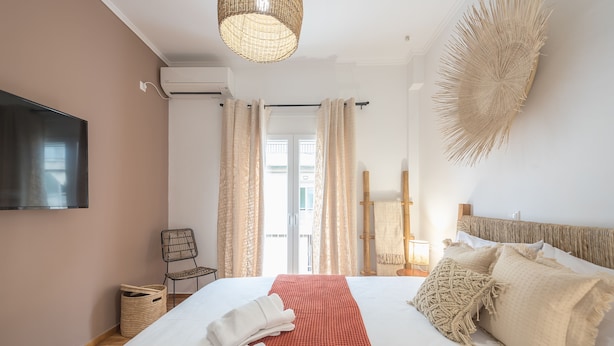 A hotel room with a bed, a TV mounted on the wall, a wicker chair, and a large woven wall decoration. - Boho Pennie Home by CloudKeys (Athens)