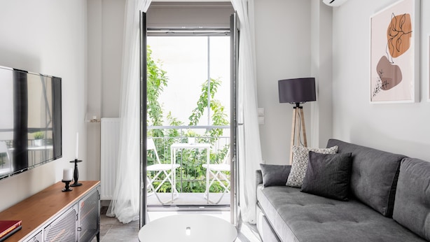 Ett modernt vardagsrum med en grå soffa, ett litet runt bord, en tv på en träkonsol och en balkong med vita gardiner.