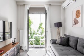 Ein modernes Wohnzimmer mit einem grauen Sofa, einem kleinen runden Tisch, einem Fernseher auf einer Holzkonsole und einem Balkon mit weißen Vorhängen.