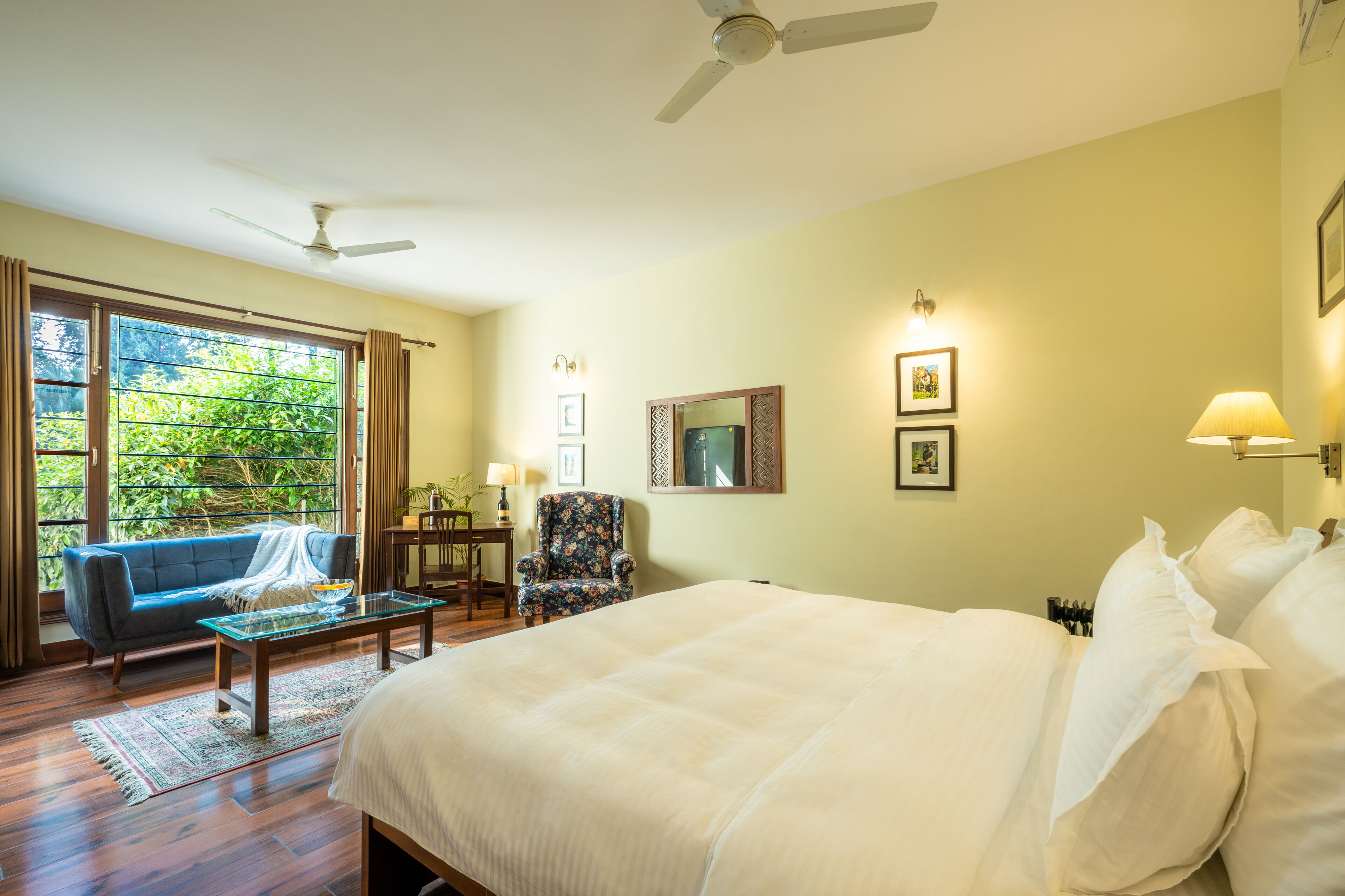 A hotel room with a large bed, a sofa, a chair, a desk, and a view of greenery through the windows.
