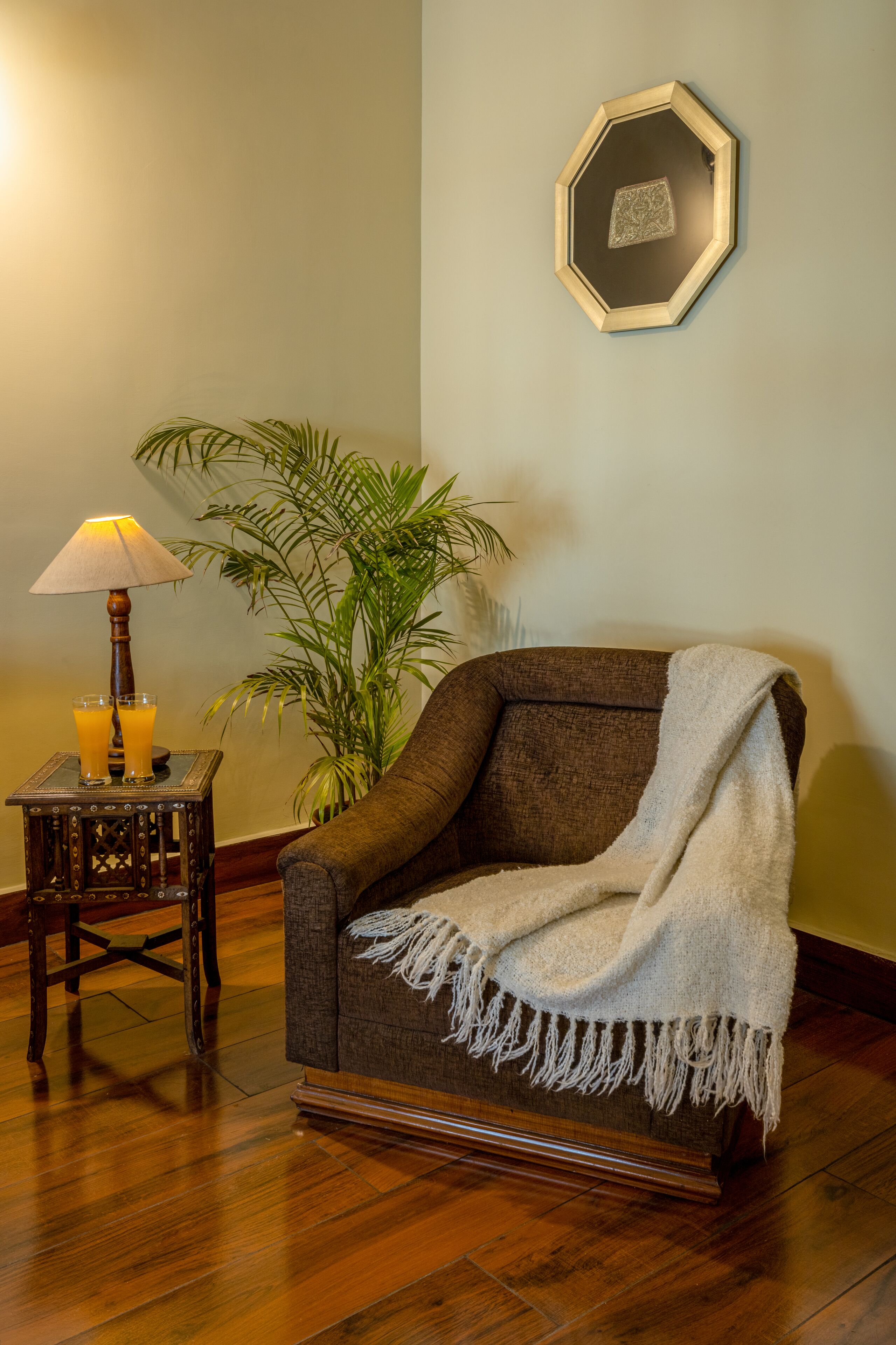Una habitación acogedora con suelo de madera, un sillón marrón con una manta blanca, una pequeña mesa de madera con una lámpara y vasos de jugo de naranja, y una planta verde grande.