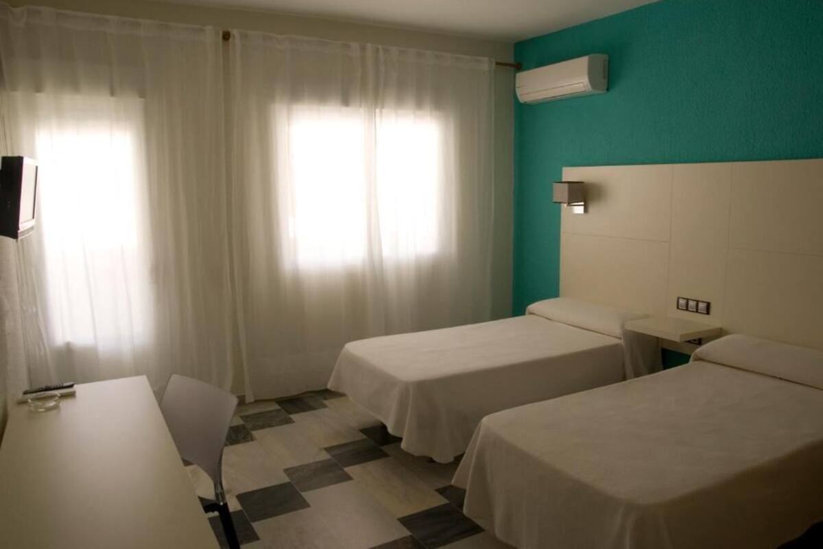 A hotel room with two single beds, a desk, a chair, a TV, and a window with curtains.