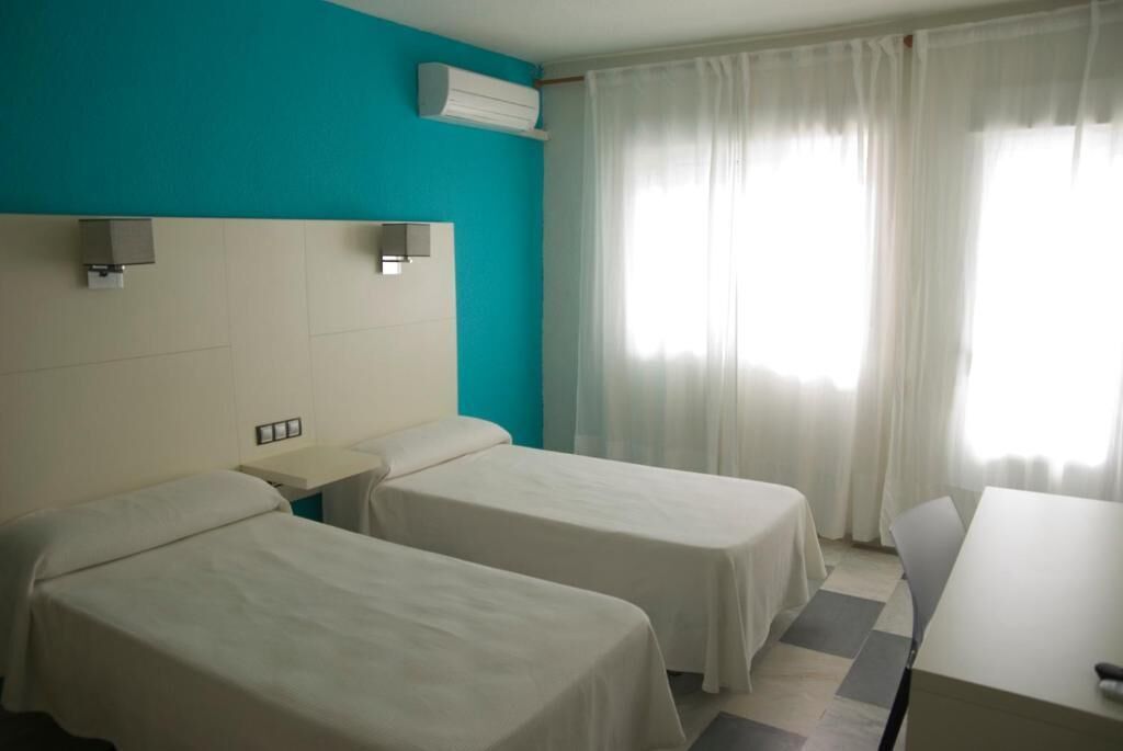 Une chambre d’hôtel avec deux lits simples, une petite table de chevet, un bureau et une fenêtre avec des rideaux.