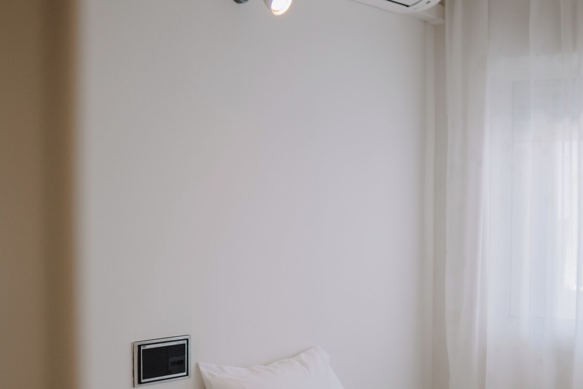 Habitación de hotel con dos camas, aire acondicionado de pared, mesita de noche con lámpara y cortinas translúcidas.