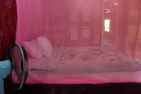 A bedroom with a bed covered by a mosquito net, a bedside fan, and a window with blinds.