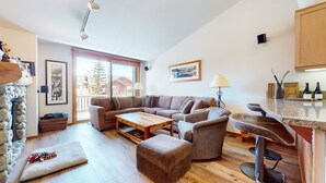 Een gezellige woonkamer met een ruime hoekbank, een houten salontafel en een comfortabele poef. Aan de ene kant is een stenen muur en aan de andere kant is een keuken met een bar.
