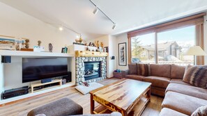 A living room with a stone fireplace, a flat-screen TV, a wooden coffee table, and a brown sofa. - Large Home, Beautiful Mountains Views, Private Washer/Dryer & 1 Car Garage (Mammoth Lakes)