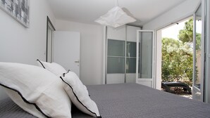 A bedroom with a bed, white pillows, a grey quilted bedspread, a white pendant light, and a sliding glass door leading to a garden. - Apartment 60 M2 with 2 Bedrooms and 2 shower rooms Sea View 300 M from the sea (Sainte-Maxime)