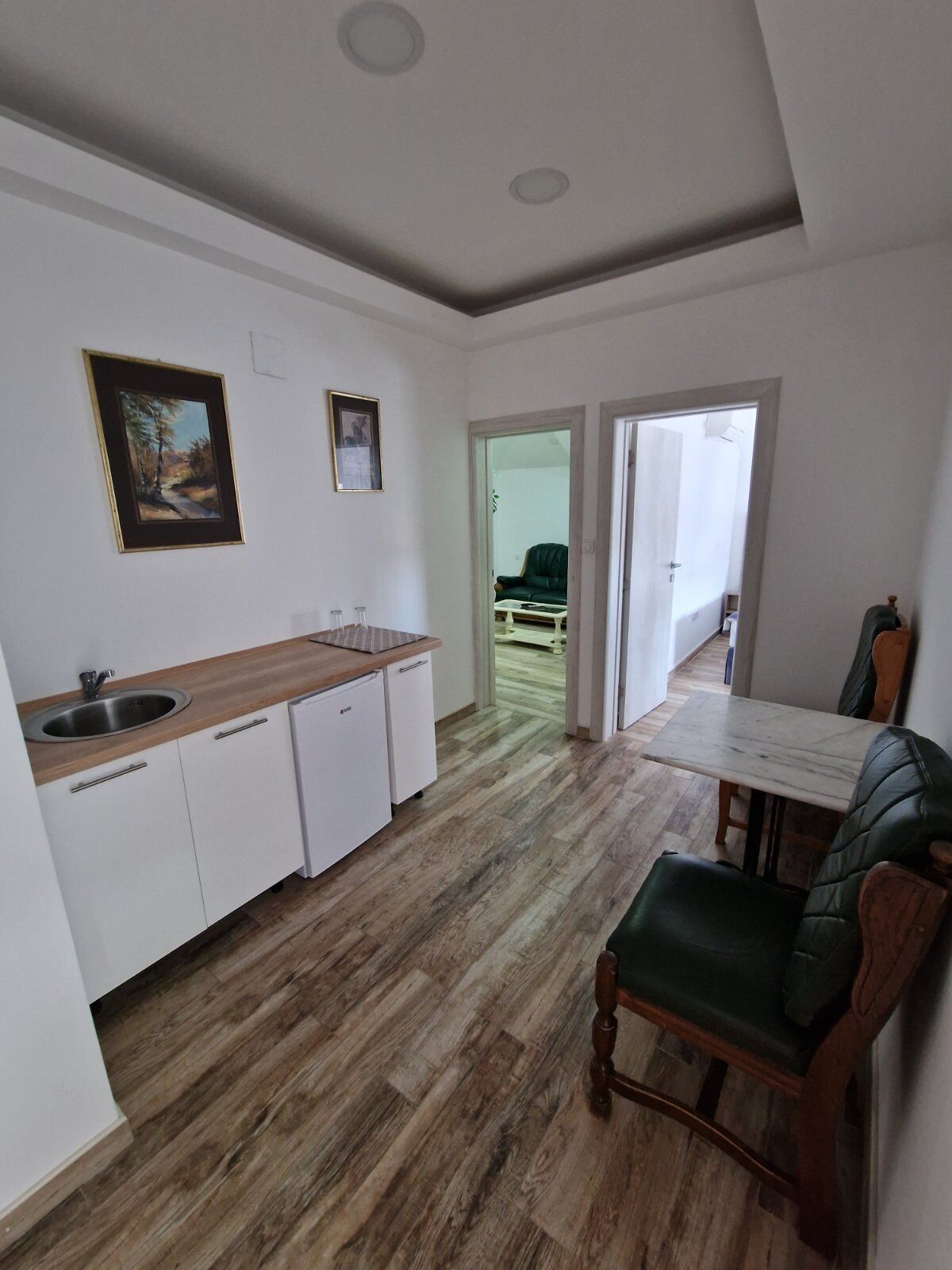 A compact kitchen with white cabinets, a wooden countertop, and a sink. There is a dining area with a table and a green upholstered chair.