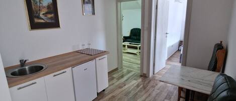 A compact kitchen with white cabinets, a wooden countertop, and a sink. There is a dining area with a table and a green upholstered chair.