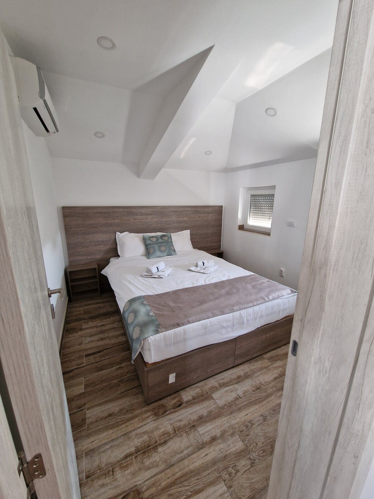 Un dormitorio con cabecera de madera, cama con ropa de cama blanca, una mesita de noche pequeña y una ventana con persianas.