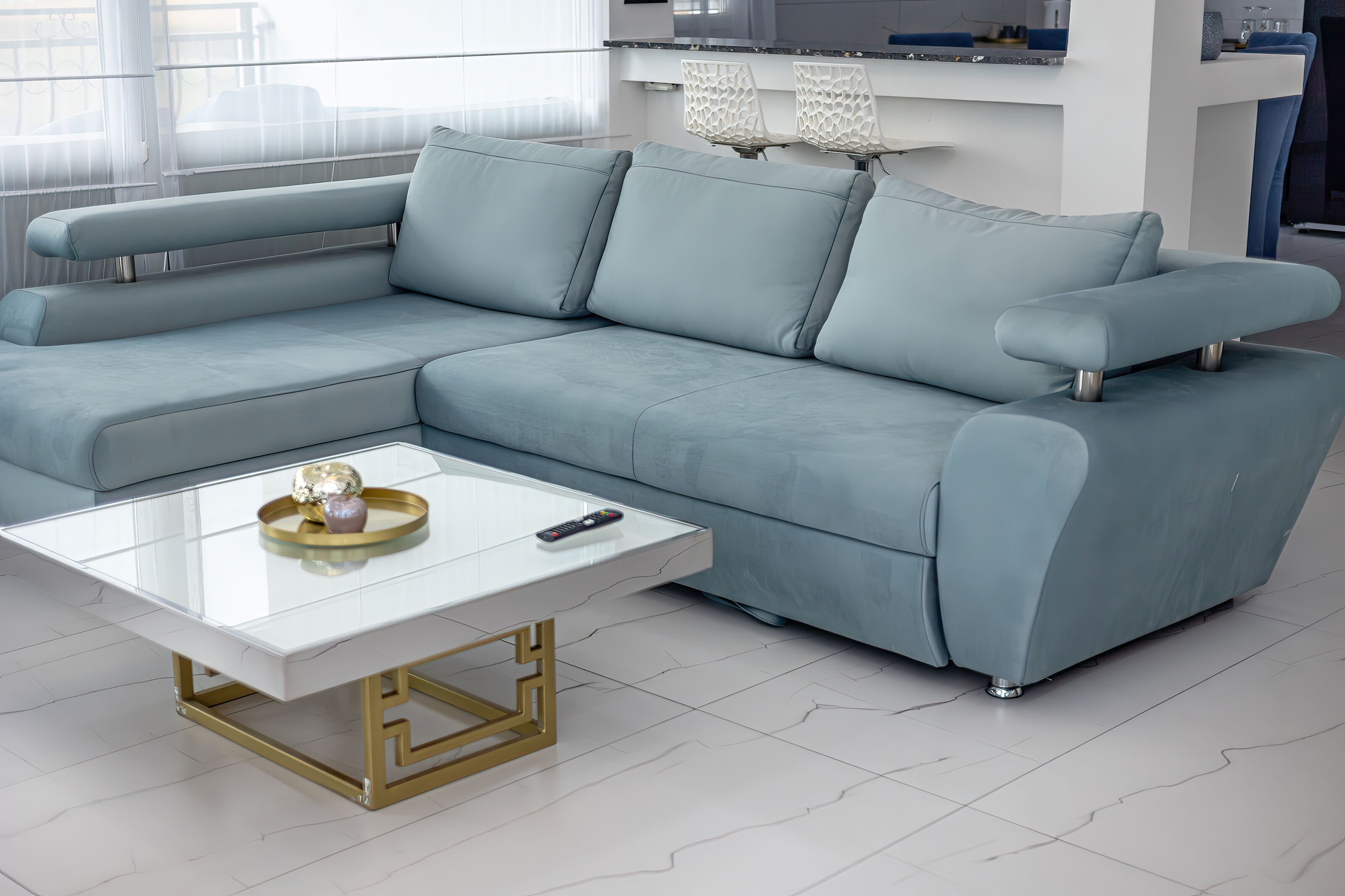 A modern living room with a light blue sectional sofa, a glass coffee table with gold legs, and a remote control on it.