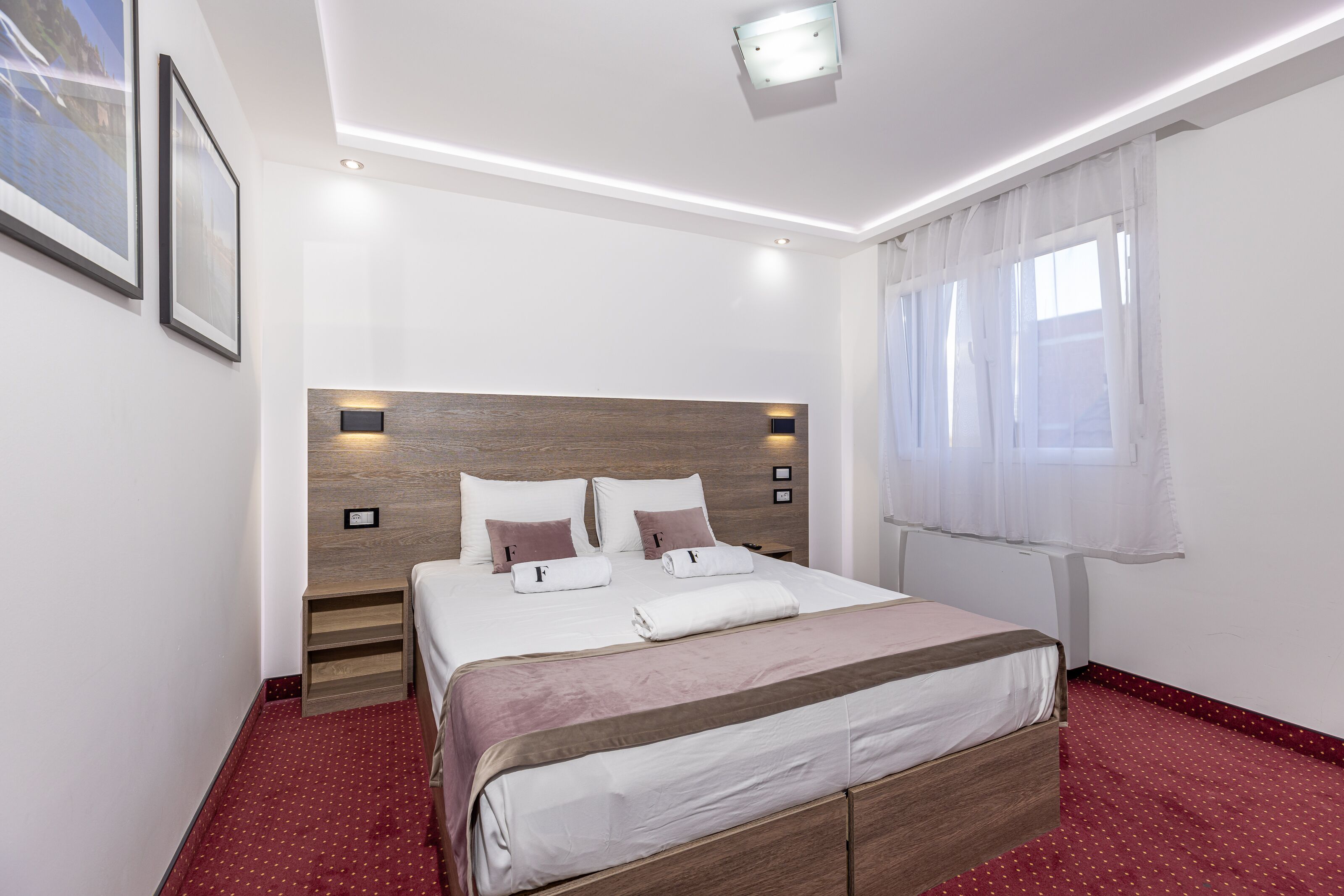 A neatly arranged bedroom with a large bed, bedside tables, a wall-mounted TV, and a window with sheer curtains.