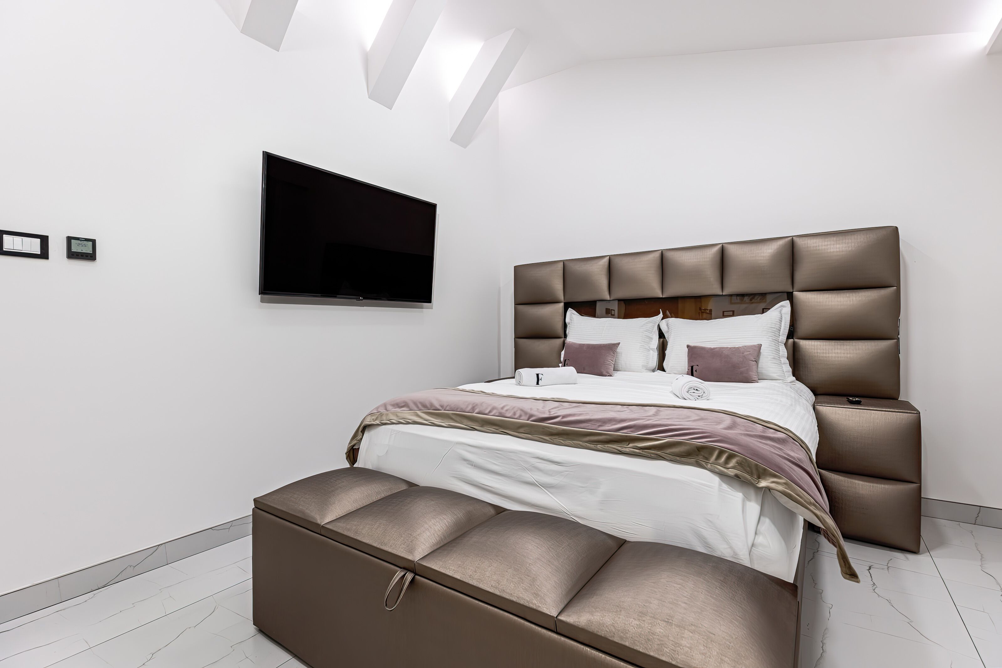 A modern bedroom with a large bed, a flat-screen TV mounted on the wall, and a built-in bench at the foot of the bed.