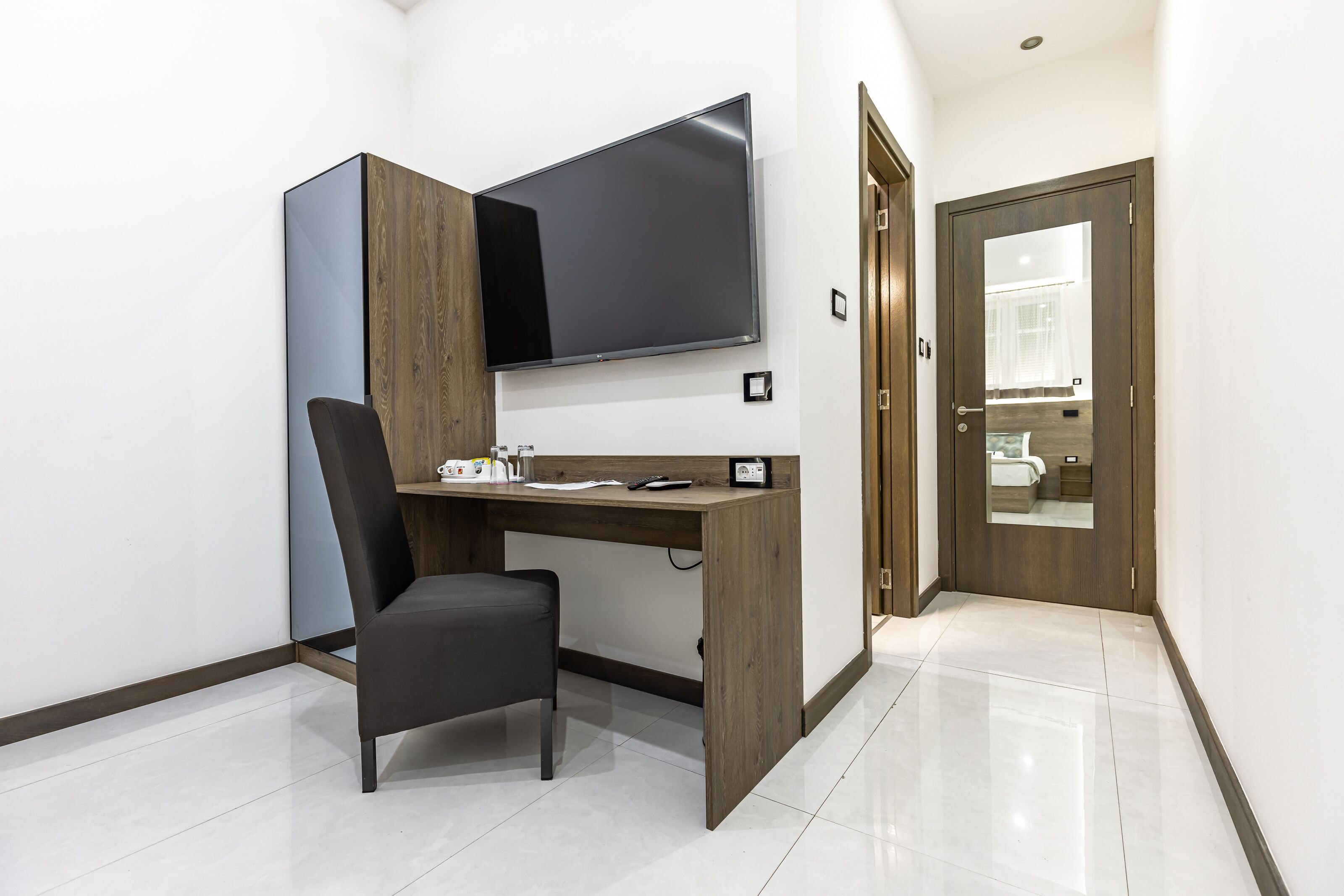 Quarto moderno com escrivaninha de madeira, cadeira preta, televisão de tela plana fixada na parede e porta de madeira que dá para outro cômodo.