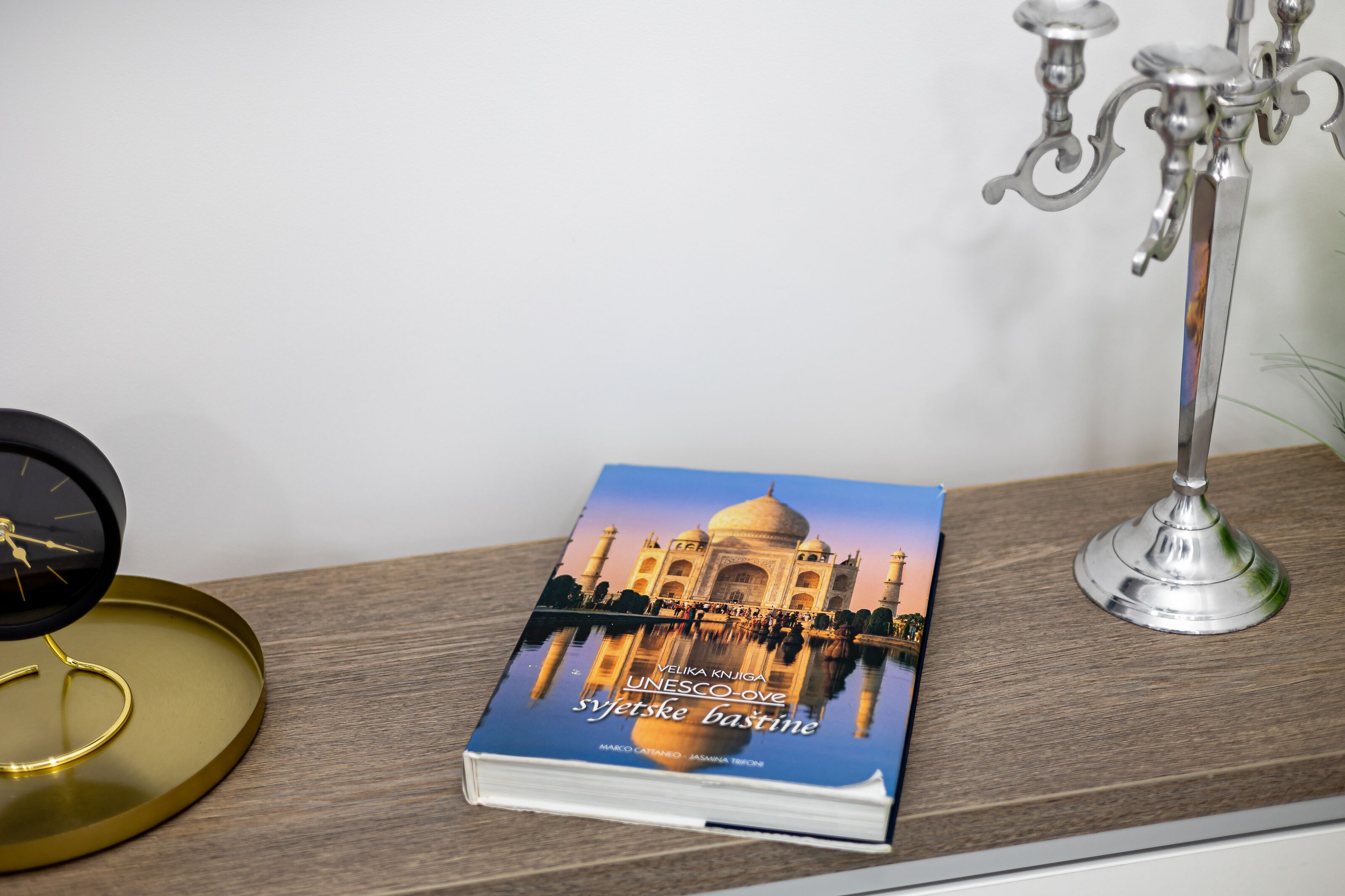 A book titled "UNESCO-ove syetiske bastine" on a wooden surface, a black clock, and a silver candelabra.