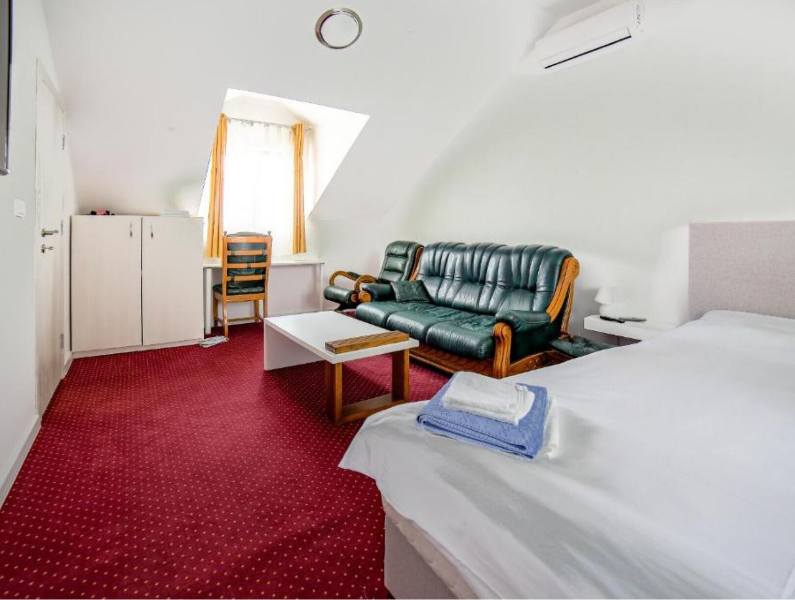 Et hotelværelse med rødt tæppe, grøn lædersofa, hvid seng og et lille bord med et blåt håndklæde.