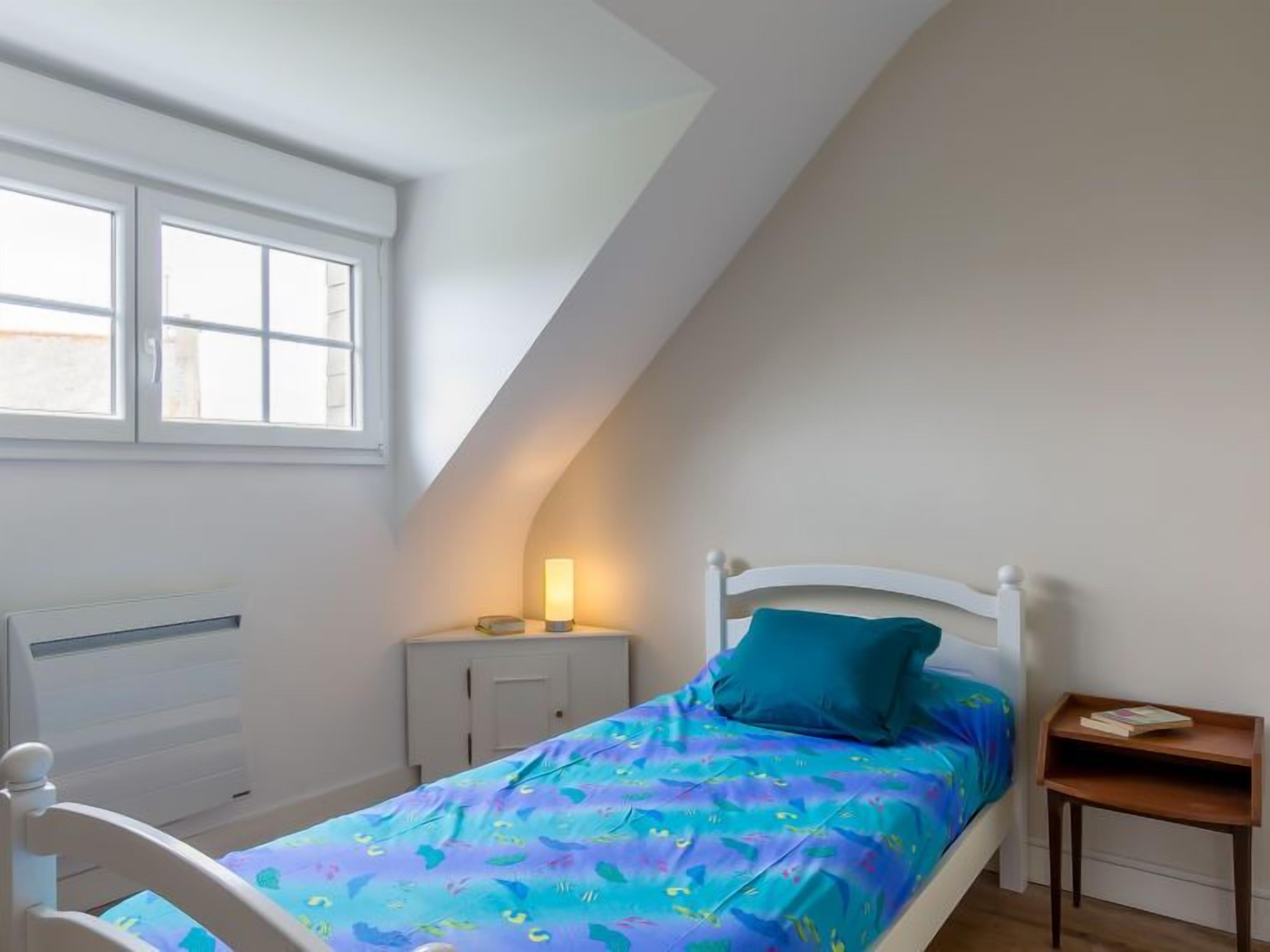 A bedroom with a sloped ceiling, a bed with a blue and purple patterned bedspread, a white headboard, a bedside table with a lamp, and a small wooden nightstand.