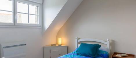 A bedroom with a sloped ceiling, a bed with a blue and purple patterned bedspread, a white headboard, a bedside table with a lamp, and a small wooden nightstand.