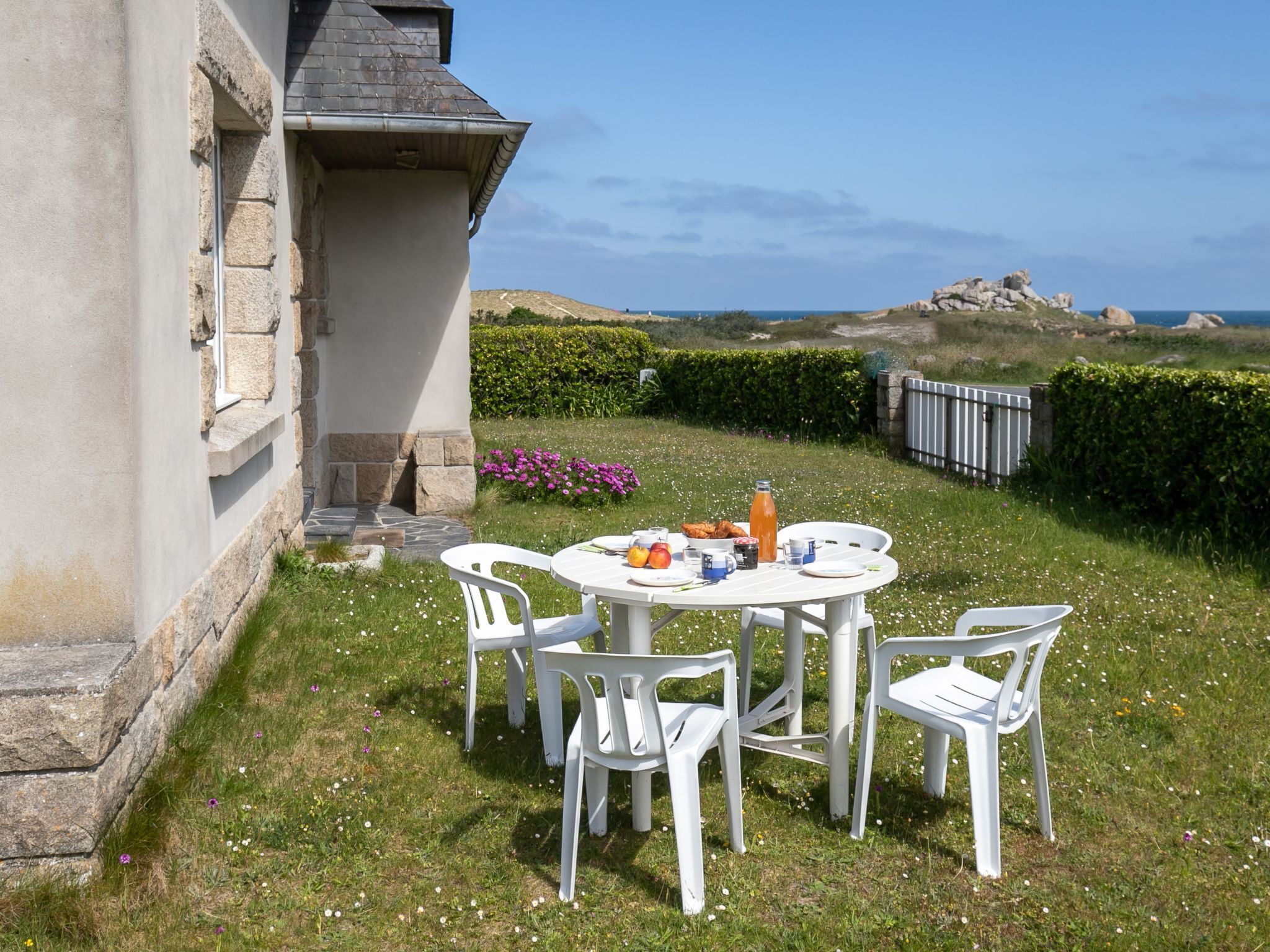 Ein runder Tisch mit weißen Stühlen steht draußen, umgeben von einer grünen Fläche mit Blumen und Blick auf Meer und Hügel im Hintergrund.